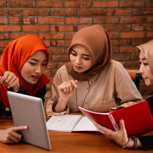 Three women reviewing documents and tablet together at wooden table against brick wall.