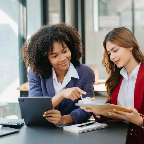 Two professional women reviewing documents and digital tablet in modern office setting with laptops and paperwork.
